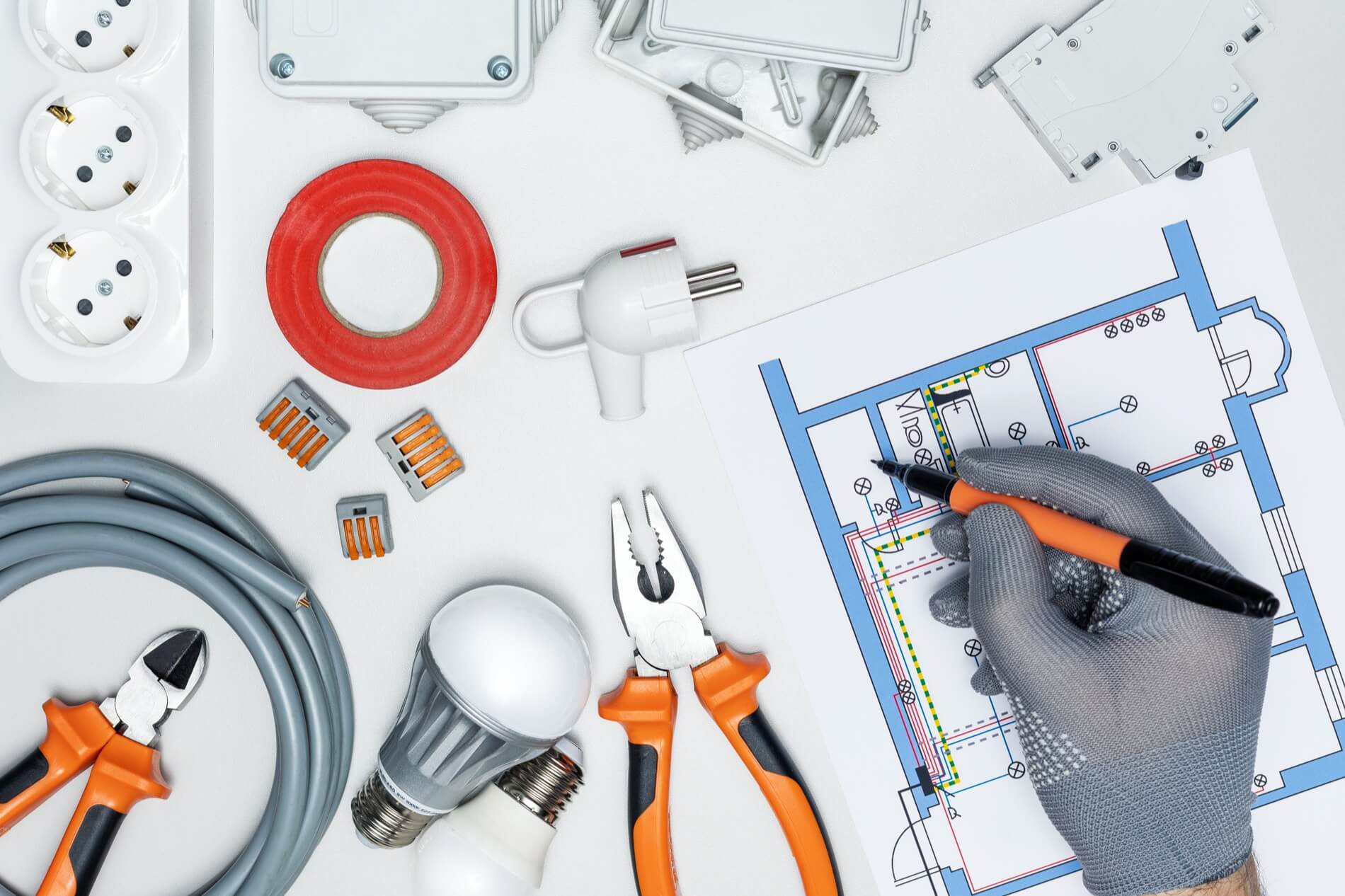 Ręka w szarej rękawicy zaznacza plan budynku wśród narzędzi i kabli, wspierając profesjonalne kosztorysowanie instalacji elektrycznych.