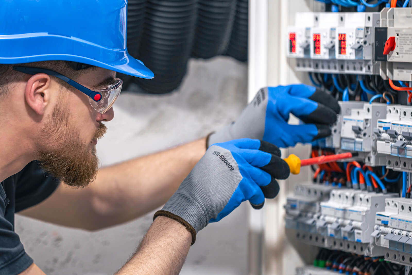 Elektryk w niebieskim kasku i rękawicach naprawia rozdzielnicę śrubokrętem, realizując projekt instalacji elektrycznej.